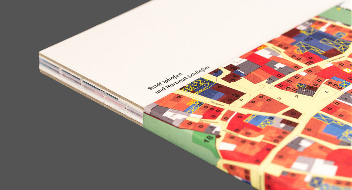 Nahaufnahme eines Buches, dessen Banderole eine detaillierte Karte in verschiedenen Farben zeigt. Der Buchrücken ist sichtbar und lässt die gebundenen Seiten erkennen. Die Herausgeber auf dem Einband sind sichtbar: „Stadt Iphofen und Hartmut Schließer”.