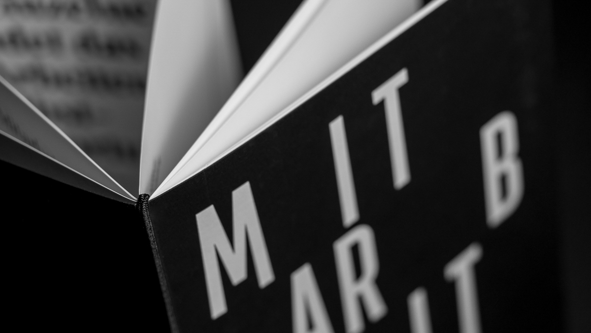 Offenes Buch in Graustufen, leicht angewinkelt, um Seiten zu zeigen, wobei der Einband einige Buchstaben von MITARBEITERFIBEL zeigt.