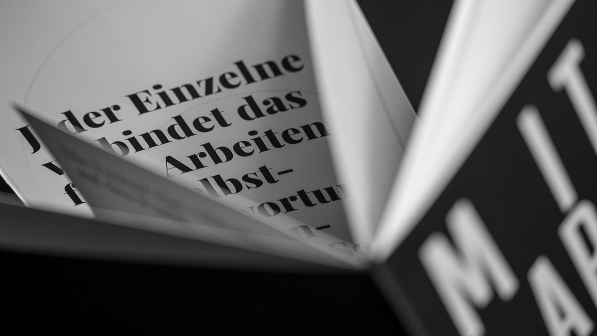 Nahaufnahme der Seiten einer Mitarbeiterfibel in Deutsch, schwarz-weiß, mit auffälligem Text im Hintergrund.